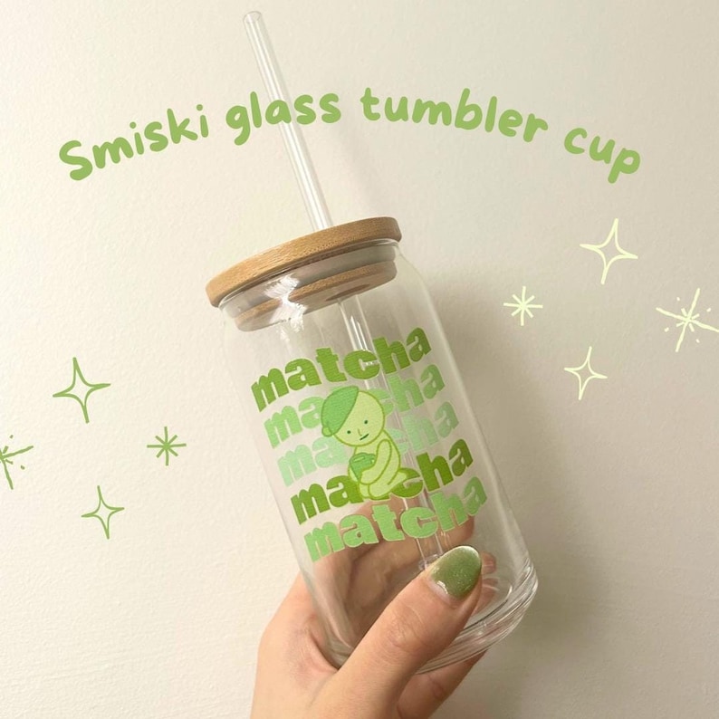 May include: A clear glass tumbler with a bamboo lid and a green and white illustration of a cartoon character holding a cup of matcha. The tumbler has the word "matcha" repeated multiple times on the side.