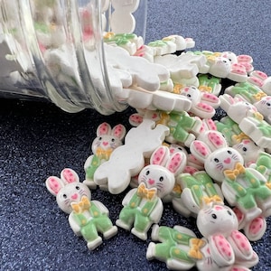 以下が含まれることがあります： 緑とピンクのアクセントが付いた白いセラミック製のウサギのチャームが入った瓶。チャームは瓶から暗い青色の表面にこぼれ落ちている。