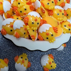 以下が含まれることがあります： 黄色と白のひよこ形のレジンカボションが入ったボウル。カボションは、卵から孵化するひよこの形をしています。ひよこの中には、完全に孵化したひよこもいれば、まだ卵の中にいるひよこもいます。