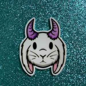 Może przedstawiać: Biały królik z kreskówki z fioletowymi rogami i różowymi uszami. Postać ma białe tło i znajduje się na turkusowym tle z brokatem.