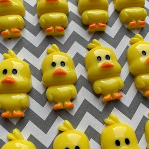 以下が含まれることがあります： 黒い目とオレンジ色のくちばしを持つ黄色いゴム製アヒルの置物のコレクション。アヒルは、グレーとホワイトのシェブロンの背景に、ずらしたパターンで配置されています。
