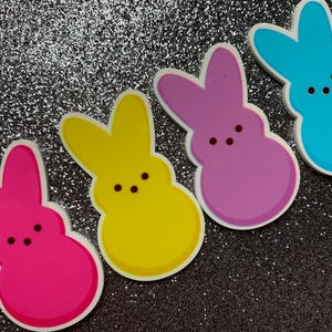 May include: Four colorful bunny-shaped marshmallow candies. The candies are pink, yellow, purple, and blue.