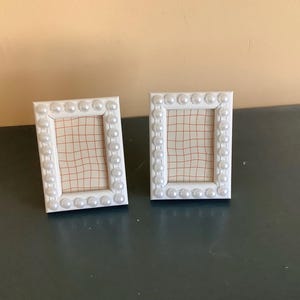 May include: Two white picture frames with a pearl-like trim. The frames are empty and have a grid pattern inside.