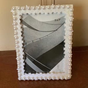 May include: White picture frame with a decorative border of faux pearls and floral lace. The frame is designed to hold a 5 x 7 inch photo. The frame is on a wooden surface.