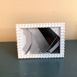 May include: A rectangular picture frame featuring a border decorated with rows of small, white, spherical beads. The frame holds a 5x7 inch display insert showing a black and white abstract architectural graphic with curved lines and geometric shapes.