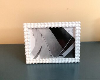 May include: A rectangular picture frame featuring a border decorated with rows of small, white, spherical beads. The frame holds a 5x7 inch display insert showing a black and white abstract architectural graphic with curved lines and geometric shapes.