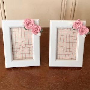 May include: Two white picture frames with pink rose embellishments. Each frame has a grid-patterned background. The frames are displayed on a brown wooden surface.
