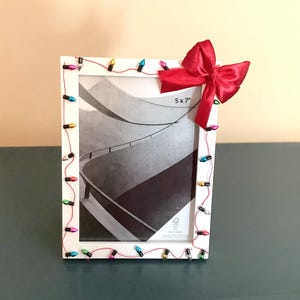 May include: White picture frame decorated with colorful Christmas lights and a red bow. The frame holds a black and white photo and is sized 5x7 inches.