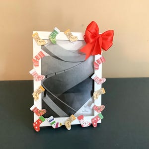May include: White picture frame decorated with small, colorful Christmas stockings. A large red bow is attached to the top right corner. The frame surrounds a black and white photograph.