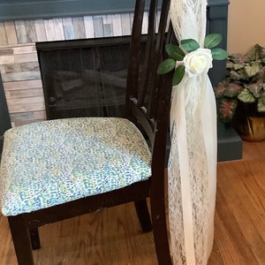 May include: A wooden chair with a patterned seat cushion and a white lace ribbon tied to the back with a white rose and green leaves.