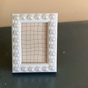 May include: A rectangular white picture frame decorated with a border of glossy white faux pearls. The frame contains an insert featuring a light-colored background with a thin, wavy orange grid pattern printed across the entire surface area.