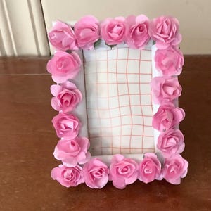 May include: A rectangular picture frame decorated with numerous small, light pink artificial roses covering the entire border. The center of the frame contains a white insert featuring a thin, light orange grid pattern printed on the surface.
