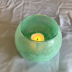 May include: A teal glass candle holder with a textured surface. The holder contains a white candle and green sprinkles.