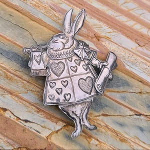May include: A silver-toned brooch depicting the White Rabbit from Alice in Wonderland. The rabbit is dressed in a waistcoat adorned with heart symbols and holds a scroll and a trumpet. The brooch is detailed with engraved lines and is set against a textured background.