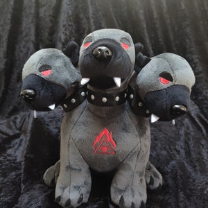 May include: A gray plush toy with three heads, each with red eyes and black noses. The toy has a studded black collar and a black and white triangle with an eye in the center on its chest.