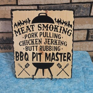 May include: A wooden BBQ Pit Master sign with black text and graphics. The sign includes the phrases "Meat Smoking," "Pork Pulling," "Chicken Jerking," and "Butt Rubbing." The sign has two small hooks below the BBQ graphic.