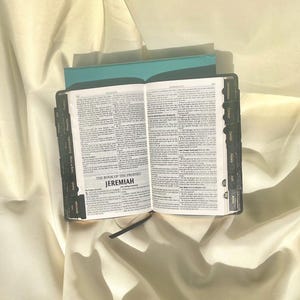 May include: An open black leather-bound book with gold lettering. The book is titled "The Book of the Prophet Jeremiah". The book has tabs for different sections of the Bible, including Song of Songs, Ecclesiastes, Proverbs, Lamentations, Ezekiel, Daniel, Hosea, Joel, and Amos.