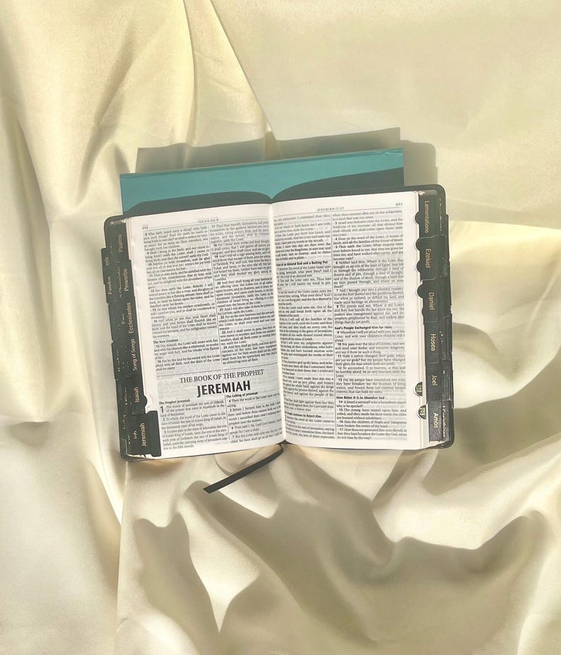 May include: An open black leather-bound book with gold lettering. The book is titled "The Book of the Prophet Jeremiah". The book has tabs for different sections of the Bible, including Song of Songs, Ecclesiastes, Proverbs, Lamentations, Ezekiel, Daniel, Hosea, Joel, and Amos.