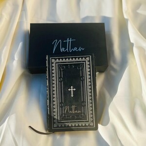 May include: A black leather-bound journal with a silver cross and ornate border design. The name "Nathan" is embossed on the front cover.