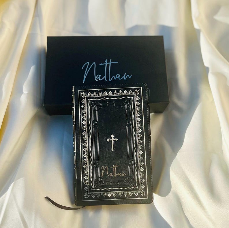 May include: A black leather-bound journal with a silver cross and ornate border design. The name "Nathan" is embossed on the front cover.
