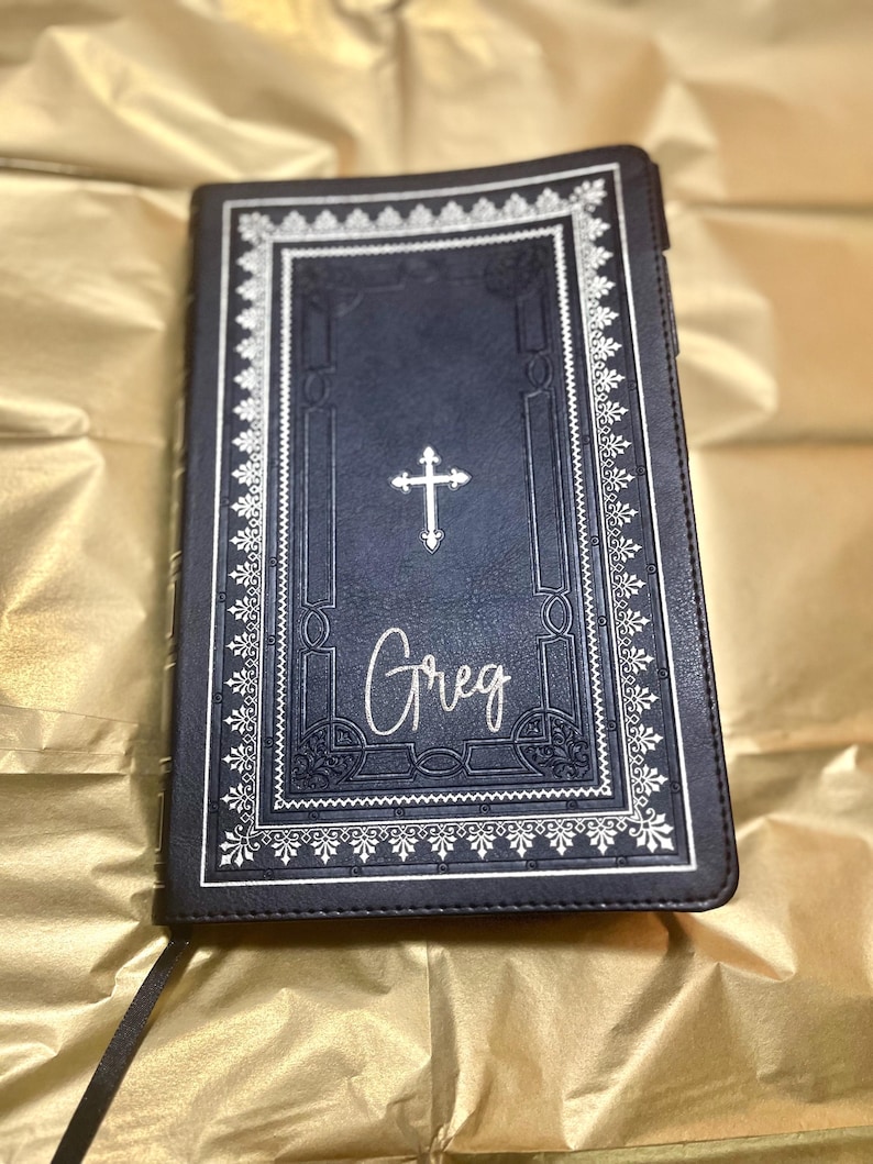 May include: A black leather-bound journal with a silver cross and the name "Greg" embossed on the front cover. The journal has a decorative silver border.