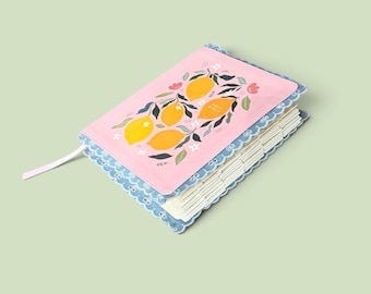 Puede incluir: Una Santa Biblia rosa con un patrón de limones y hojas en la portada, con un borde de encaje festoneado azul. El libro incluye páginas con pestañas blancas y un marcador de cinta blanca fina que sobresale por el lateral de la cubierta.