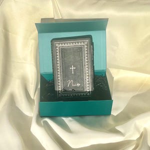 May include: A teal gift box with a black and silver book inside. The book has a cross and the name "Nico" on the cover.