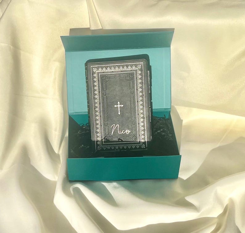 May include: A teal gift box with a black and silver book inside. The book has a cross and the name "Nico" on the cover.