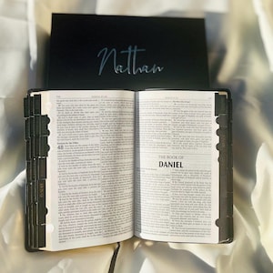 May include: An open black leather-bound Bible with gold-toned lettering on the spine. The Bible is open to the Book of Daniel, with the title "The Book of Daniel" printed in bold, black letters. The pages are filled with text in black ink. The Bible has a black bookmark with a silver inscription that reads "Nathan".