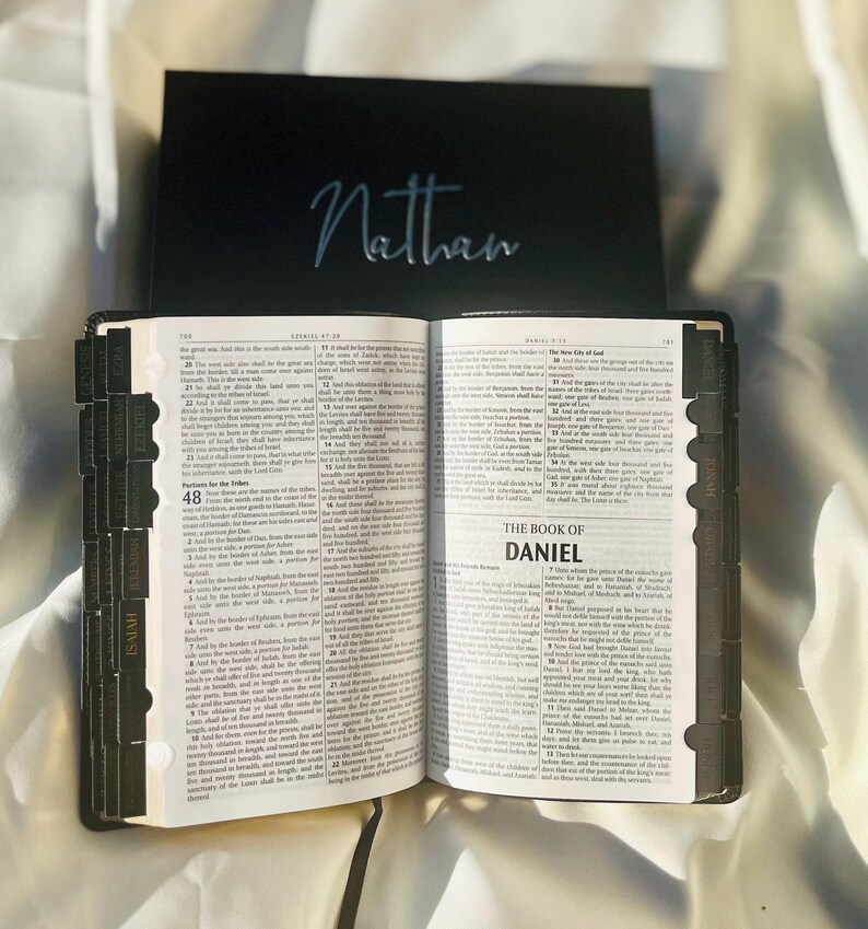 May include: An open black leather-bound Bible with gold-toned lettering on the spine. The Bible is open to the Book of Daniel, with the title "The Book of Daniel" printed in bold, black letters. The pages are filled with text in black ink. The Bible has a black bookmark with a silver inscription that reads "Nathan".