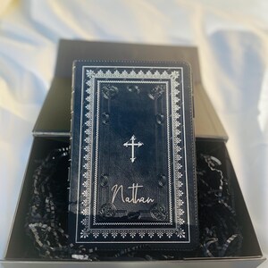 May include: A black leather-bound journal with a silver cross and the name "Nathan" embossed on the front cover. The journal is in a black gift box with black shredded paper.