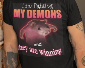 Puede incluir: Camiseta negra con un gráfico rosa y blanco de una rata con el texto "i am fighting MY DEMONS and they are winning".
