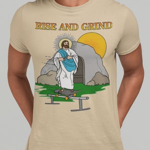 Op de afbeelding: Beige T-shirt met een cartoon illustratie van Jezus die aan het skateboarden is. De afbeelding bevat een zon, een graf en de tekst "RISE AND GRIND" in gouden letters. Het ontwerp staat op de voorkant van het shirt.