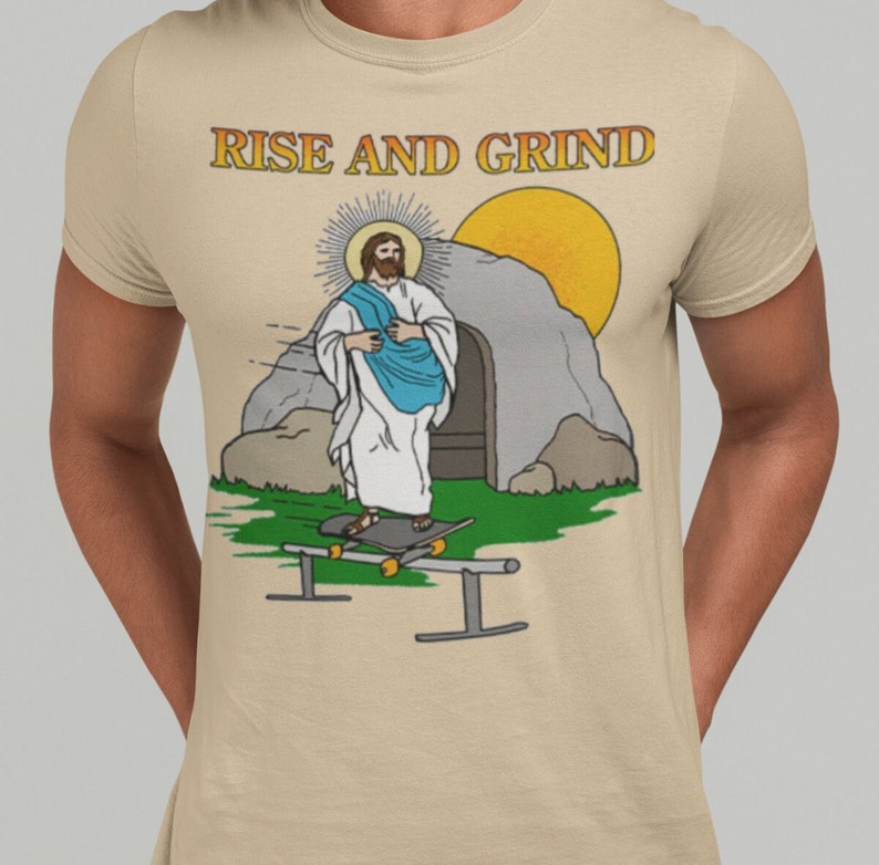 Op de afbeelding: Beige T-shirt met een cartoon illustratie van Jezus die aan het skateboarden is. De afbeelding bevat een zon, een graf en de tekst "RISE AND GRIND" in gouden letters. Het ontwerp staat op de voorkant van het shirt.