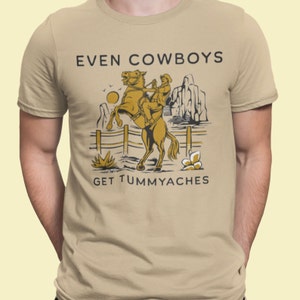May include: A tan t-shirt with a graphic of a cowboy riding a rearing horse. The text "EVEN COWBOYS GET TUMMYACHES" is printed in black on the shirt.