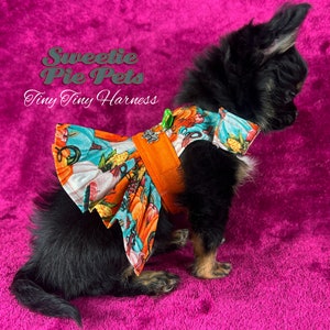 May include: A small black and brown chihuahua wearing a bright orange harness with a pumpkin and floral print dress. The text "Sweetie Pie Pets Tiny Tiny Harness" is on a pink background.