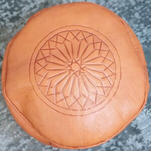 May include: Round, tan leather ottoman with a geometric starburst pattern embossed on the top. The ottoman is a warm, earthy tone and appears to be made of soft leather. The design is centered on the top surface.