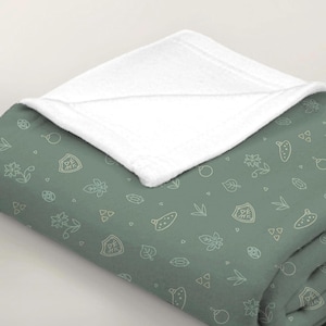 Könnte beinhalten: Eine salbeigrüne Wohndecke mit weißer Fleece-Unterseite und einem wiederkehrenden Muster aus hellen Strichzeichnungen, darunter Schilde, Okarinas, Blumen, Blätter und kleine geometrische Formen, die über den Stoff verteilt sind.