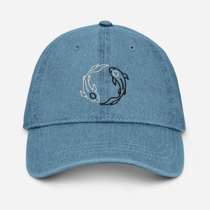 May include: A blue denim baseball cap with a black and white yin yang symbol featuring two fish. The fish are outlined in black and the yin yang symbol is filled in with black and white.