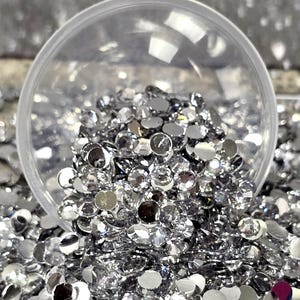 May include: A clear plastic scoop filled with silver rhinestones. The scoop is sitting on a pile of silver rhinestones.