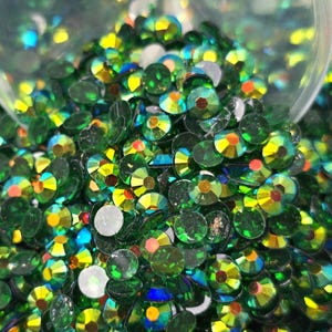 May include: A close-up of a large pile of green iridescent rhinestones. The rhinestones are round and have a faceted surface. They are scattered in a random pattern.