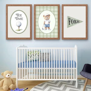 May include: Three framed golf-themed prints. One print says "Tee Time" with a golf ball illustration. Another features a teddy bear in golf attire. The third print says "Fore" on a pennant. The prints are in a nursery setting.
