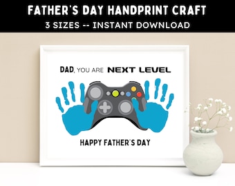 May include: A white frame with a printable Father's Day craft. The craft features a blue handprint on each side of a black video game controller. The text reads "Dad, you are next level. Happy Father's Day."