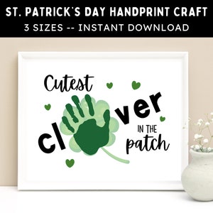 以下が含まれることがあります： 白枠のプリントで、黒と緑で「Cutest clover in the patch」と書かれています。緑の手形が中央にあり、クローバーの形をしています。小さな緑色のハートがテキストの周りに散らばっています。