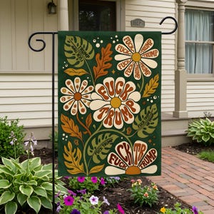 Può includere: Una bandiera da giardino rettangolare con sfondo verde scuro e un design floreale in stile retrò. I fiori hanno petali bianchi e crema con centri arancioni. Il testo FUCK TRUMP è stampato ripetutamente sui petali e sulle foglie.