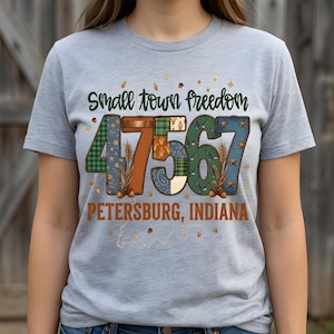 May include: A pale grey t-shirt featuring the words "Small town freedom" in green script. The number "47567" is in a patchwork design, with "Petersburg, Indiana" below in orange text.