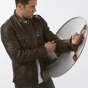 May include: A brown leather jacket with a zipper closure and multiple pockets. The person is holding a silver and red shield with leather straps. The shield is approximately 24 inches in diameter.