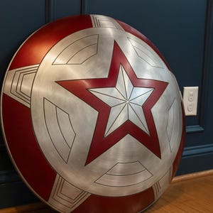 May include: A metallic silver and red shield with a large red star in the center. The shield has a circular design with red borders and geometric patterns. It is leaning against a dark blue wall on a wooden floor.