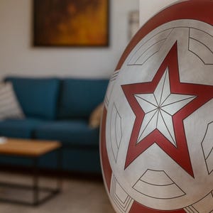 May include: A silver and red shield with a large red star in the center. The shield has a circular design with red and silver segments. The shield is leaning against a white wall, with a blurred living room in the background.