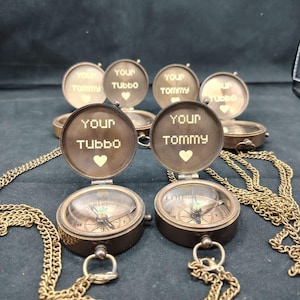 May include: Two antique brass compass necklaces with chains. The compass faces are open and feature engraved messages that say "Your Tommy" and "Your Tubbo" with a heart symbol.
